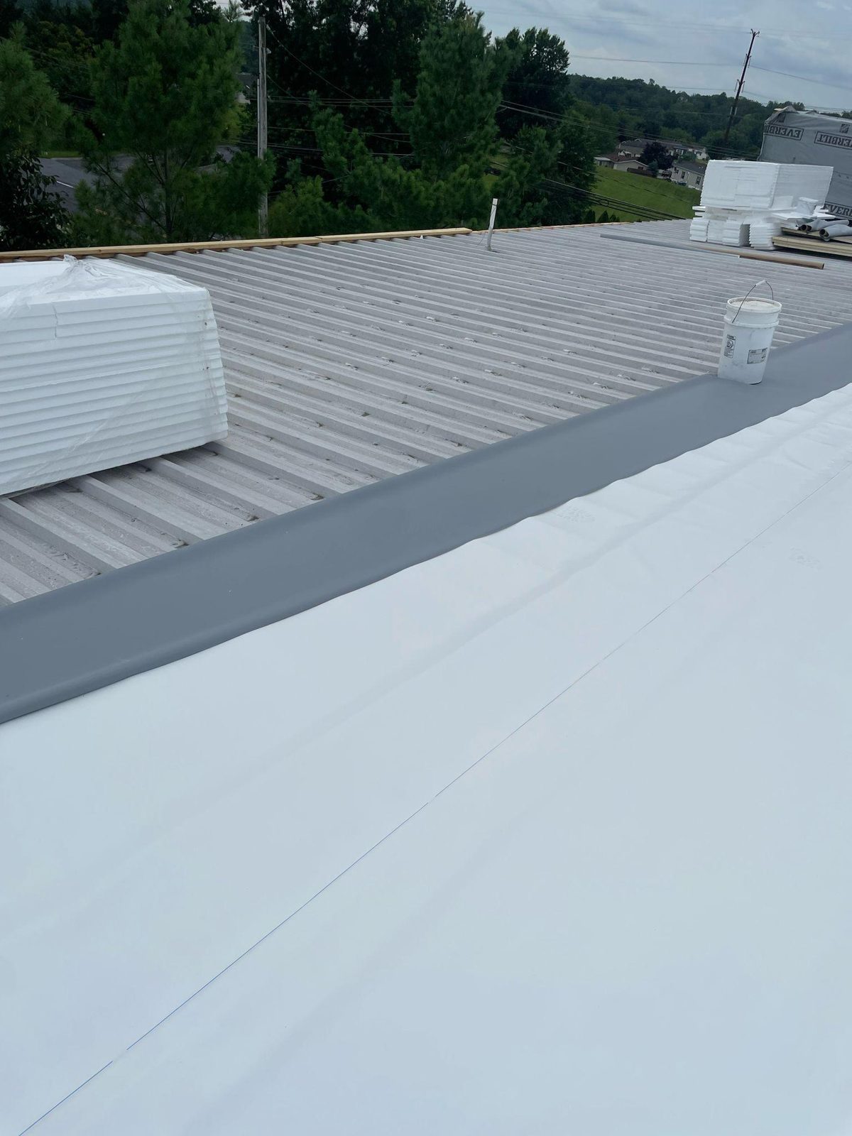A white TPO membrane being installed over corrugated commercial metal panels, with stacks of foam insulation board on the roof